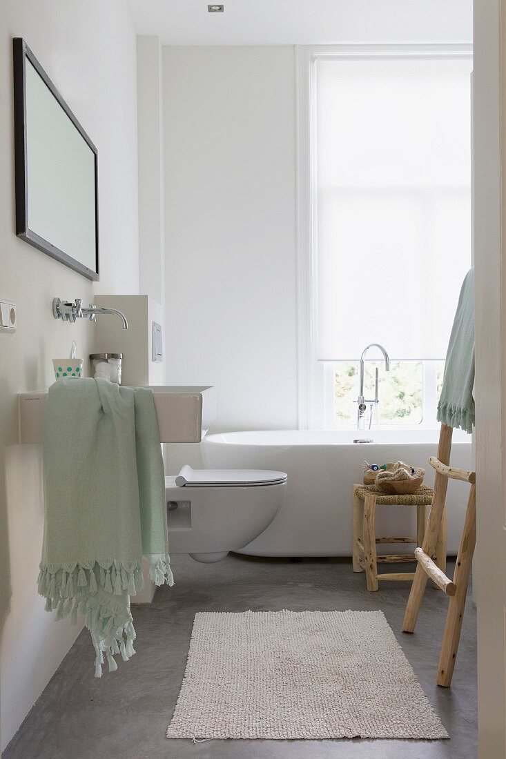 Modern bathroom with designer bathtub below window; stool and ladder-style towel rack made from plain wood
