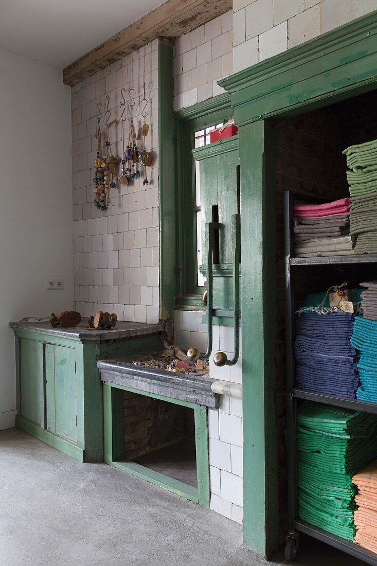Alte, grün lackierte Holzeinbauten in gefliester Wand mit Ketten-Deko; farbig sortierte, gestapelte Tücher in offenem Schrank