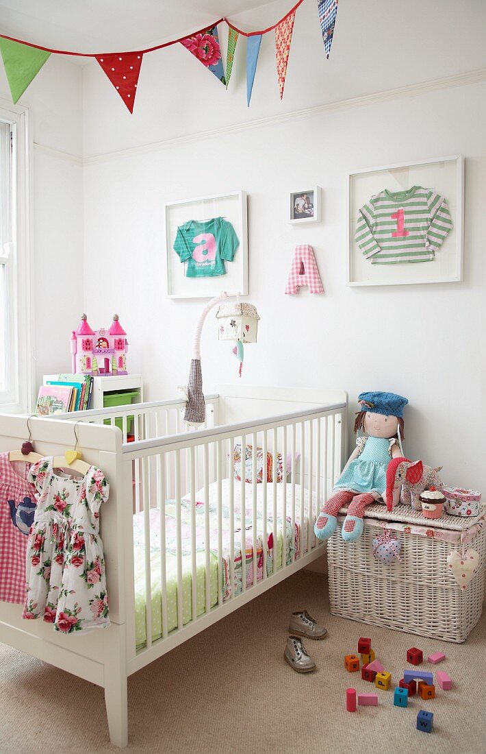 Gitterbett und weiße Rattantruhe, Puppe und Spielzeug im Kinderzimmer, an Wand aufgehängte Bilderrahmen mit Kinderkleidung