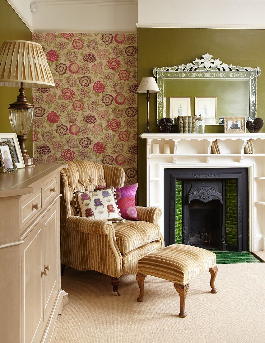 Armchair with footstool against floral wallpaper and open fireplace with shelves integrated into surround in traditional living room