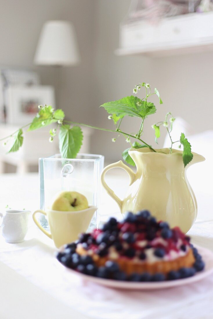 Berry tart on plate in front of lemon balm in large jug and apple in small jug