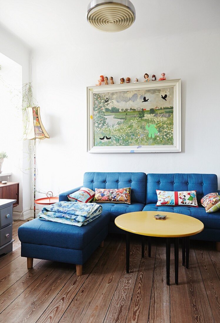 Runder Couchtisch mit gelber Tischplatte und blaues Polstersofa mit passendem Fussschemel in schlichtem Wohnzimmer, an Wand Gemälde