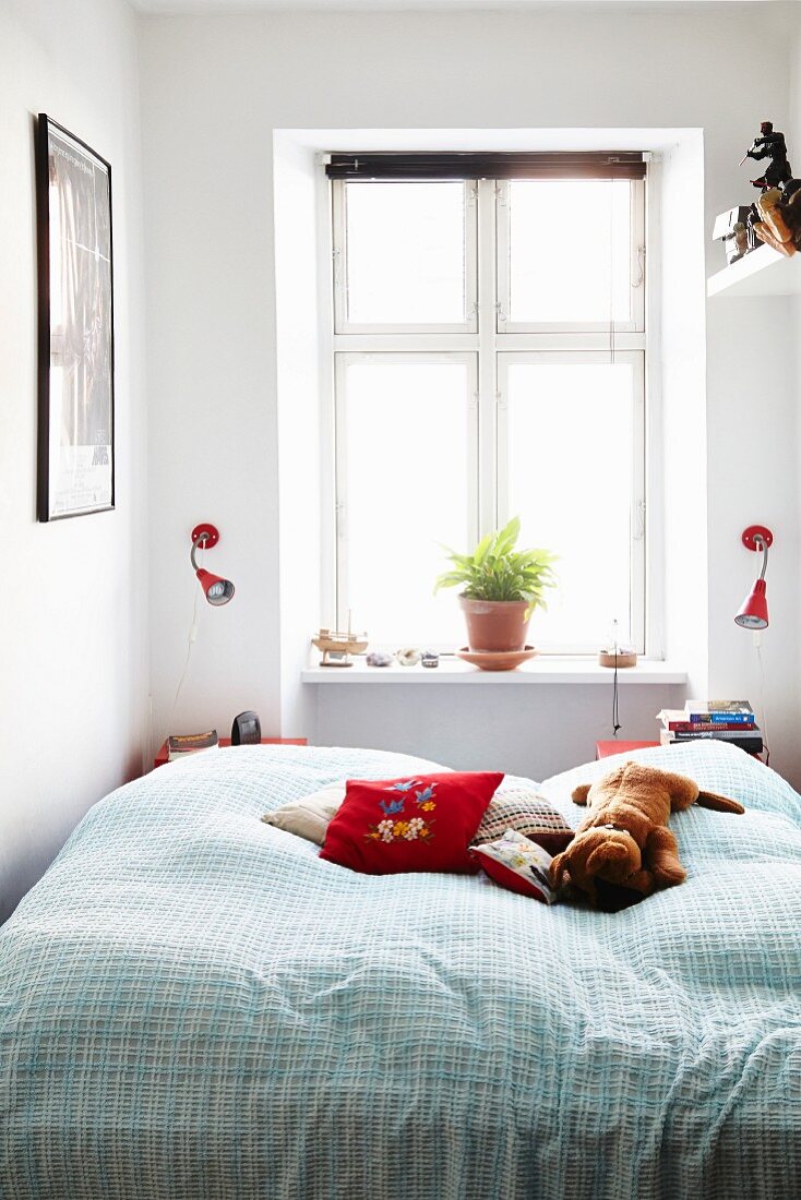 Doppelbett mit Kissen und Stofftier vor Fenster, Blumentopf auf Fensterbank