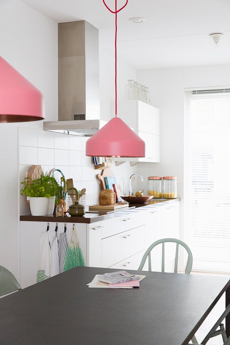 Pendant lamps with pink lampshades above black metal dining table; kitchen counter against white-tiled wall in background