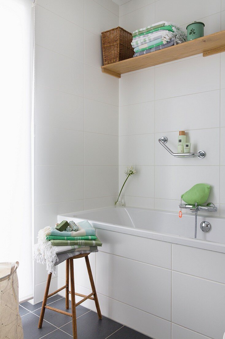 Toiletries and towels on wooden stool next to bathtub with white-tiled surround in corner of bathroom