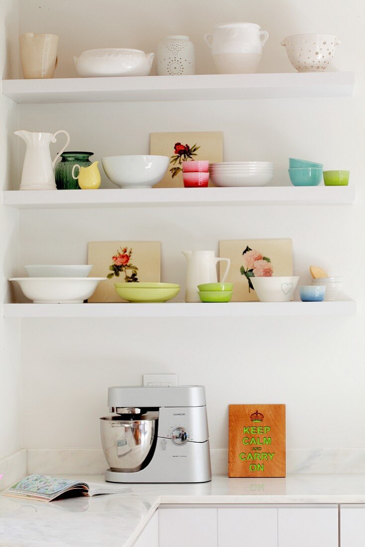 Weisses und pastellfarbenes nostalgisches Geschirr auf Regalböden an weisser Küchenwand mit Küchenmaschine auf Marmor-Küchenarbeitsplatte