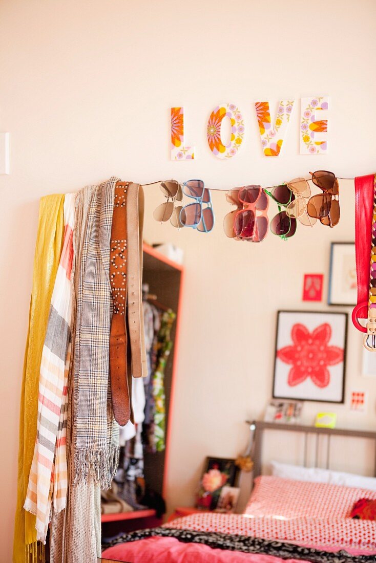 Scarves, belts and sunglasses hanging on mirror reflecting a bed with red bed linen and ornaments