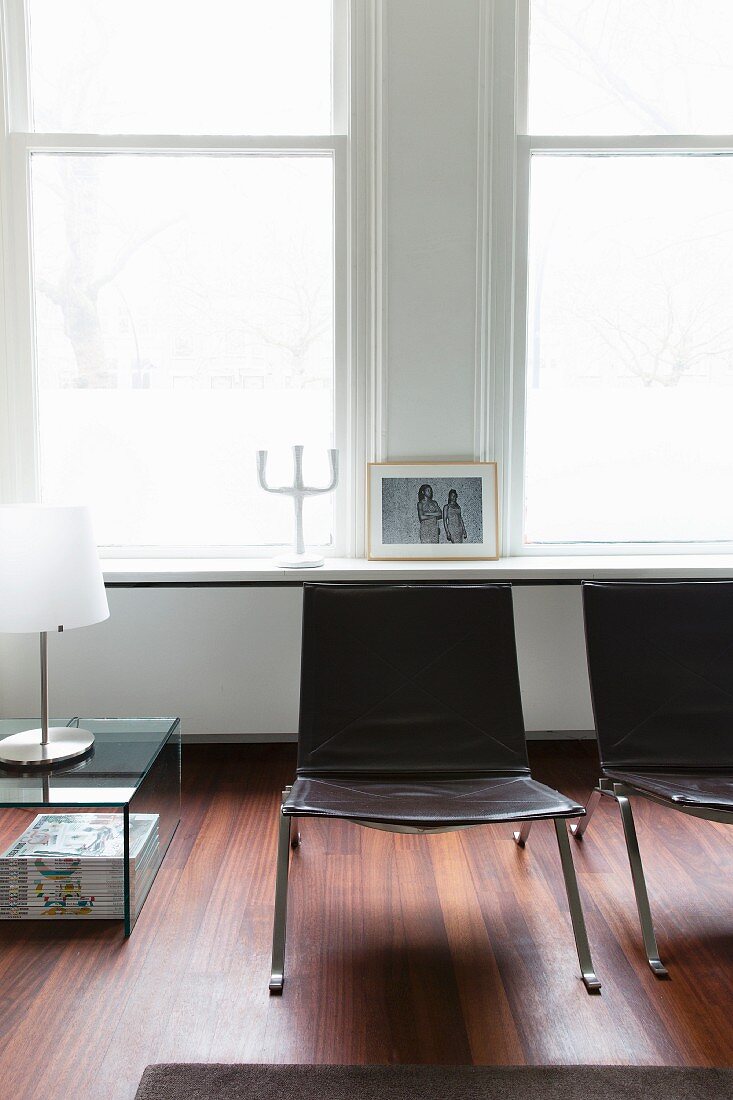 Stuhl aus Metall und Leder vor Altbaufenstern; daneben ein Glastisch mit Tischleuchte