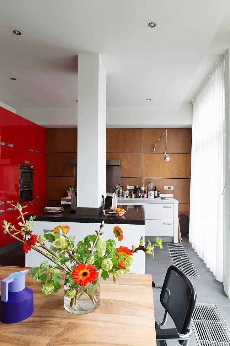 Blick von Essplatz auf Designerküche mit Kücheninsel um weiße Mauerstütze, seitlich rote Einbaufront