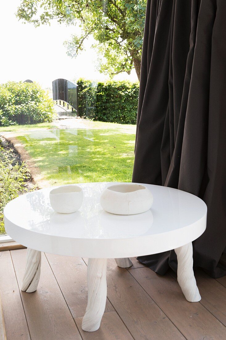 Weisser Bodentisch mit hochglänzender Platte und rustikalen Füssen vor Glasfront, Blick in sommerlichen Garten