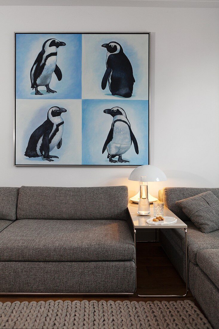 Schmaler Beistelltisch mit Retro Tischleuchte neben graumelierter Couch, oberhalb an Wand Aufnahmen von Pinguinen