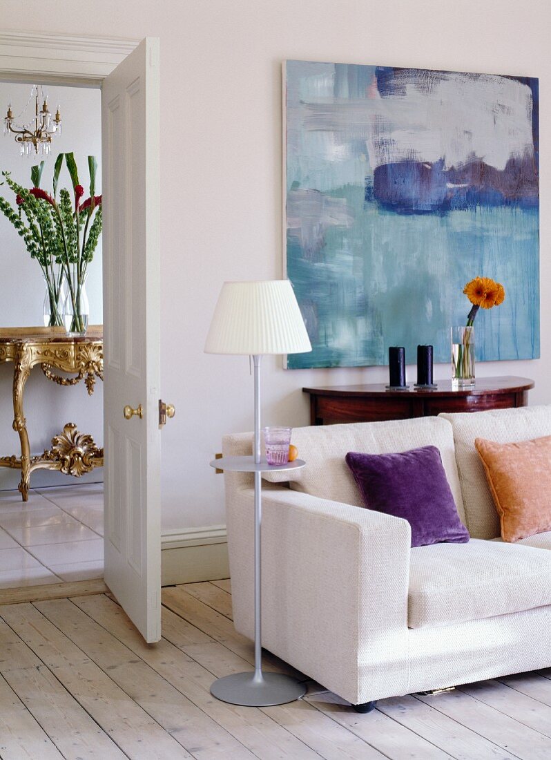 Sofa, standard lamp and console table below modern artwork; view of antique console table in hallway through open door