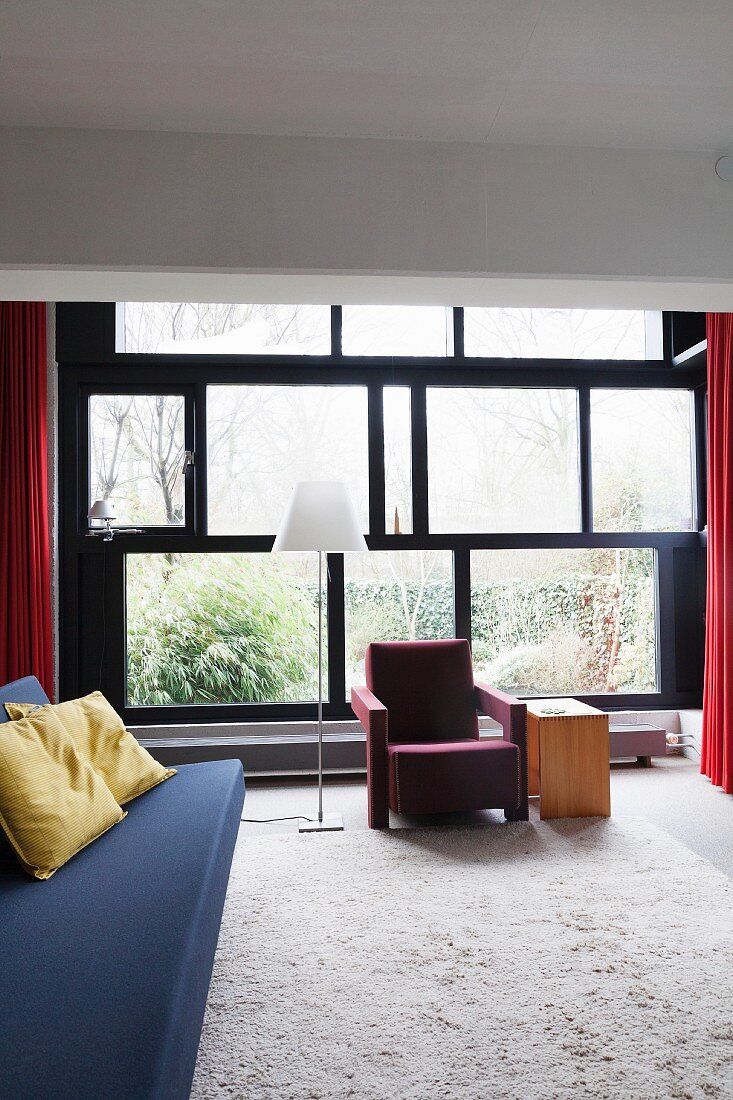 Blaue Couch und roter Sessel neben Stehleuchte, vor Fensterfront im Mondrian Stil