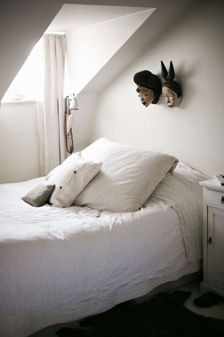 Schlichtes Bett mit Kissen und weisser Bettwäsche, unter Gaube mit Fenster, vor Wand mit aufgehängten Masken