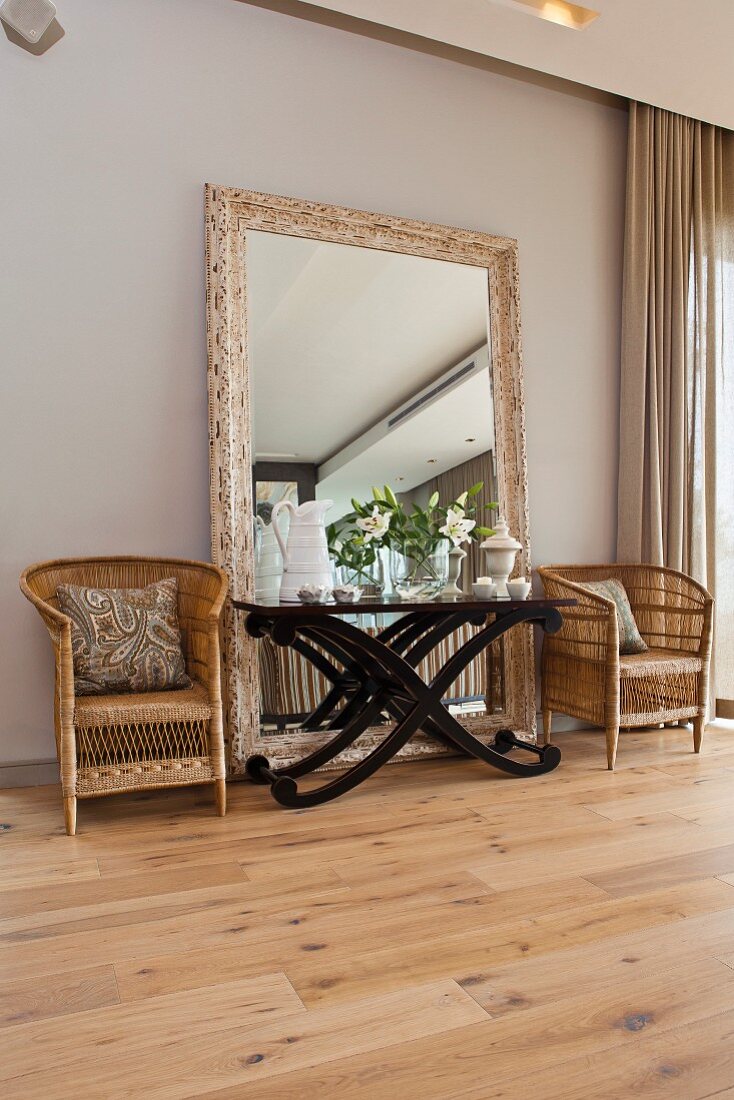 Side table with curved wooden frame in front of large free-standing mirror flanked by rattan armchairs on rustic wooden floor