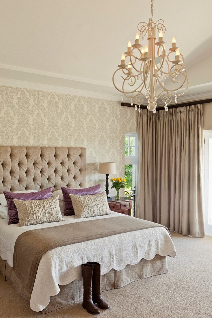 Double bed with scatter cushions and button-tufted headboard in traditional bedroom with brocade wallpaper and elegant chandelier