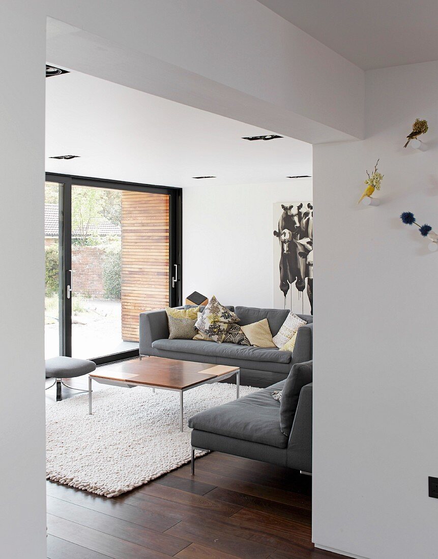 Blick durch breiten Durchgang auf graue Sofakombination und minimalistischen Couchtisch in modernem Wohnzimmer mit Gartenblick
