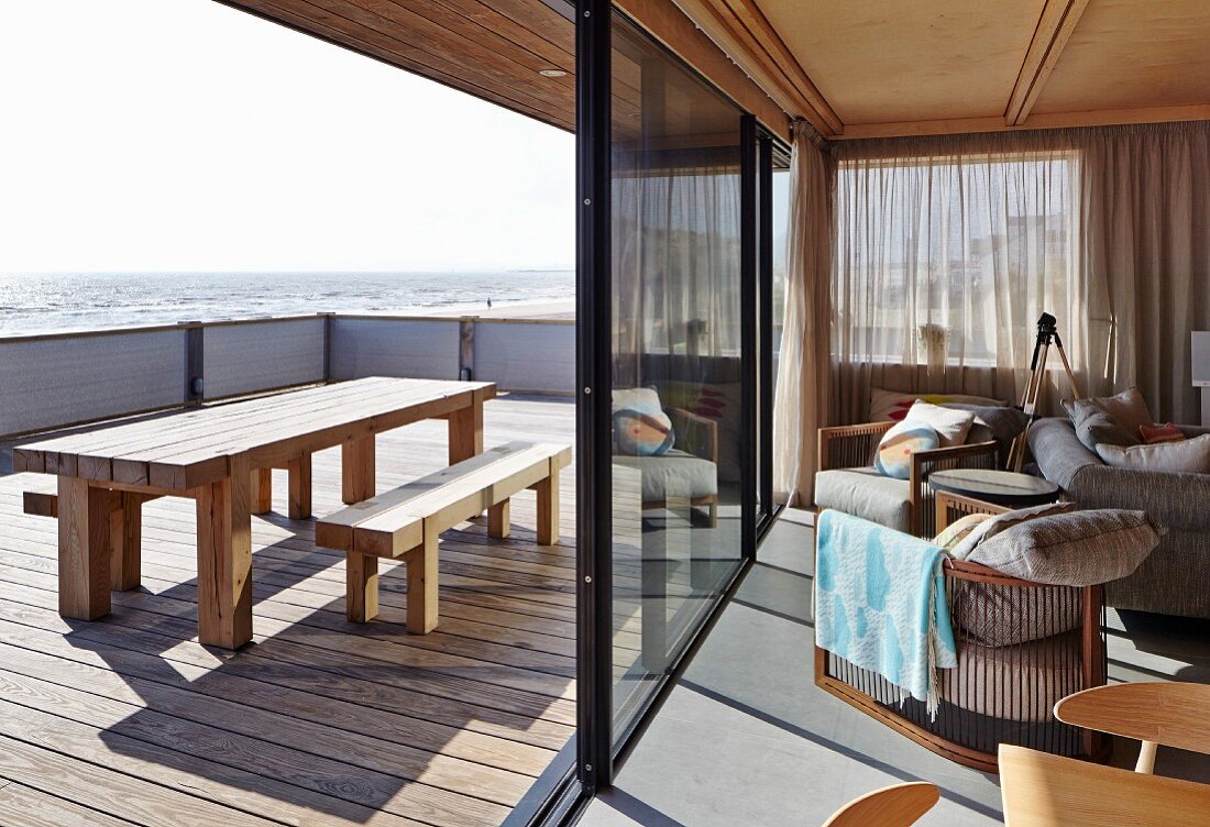Blick von Wohnzimmer über Terrasse, mit Tisch und Bank auf Holzdielenboden, auf das Meer