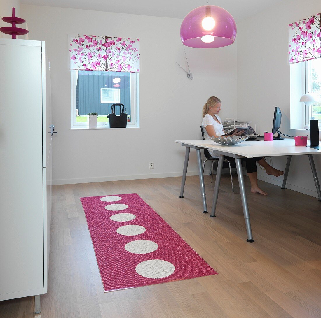 Home-Office mit Doppeltisch und Aktenschrank in schlichtem Weiß, dazu ein gepunkteter Teppichläufer, eine Plexi-Leuchte und Blumenrollos in Pink