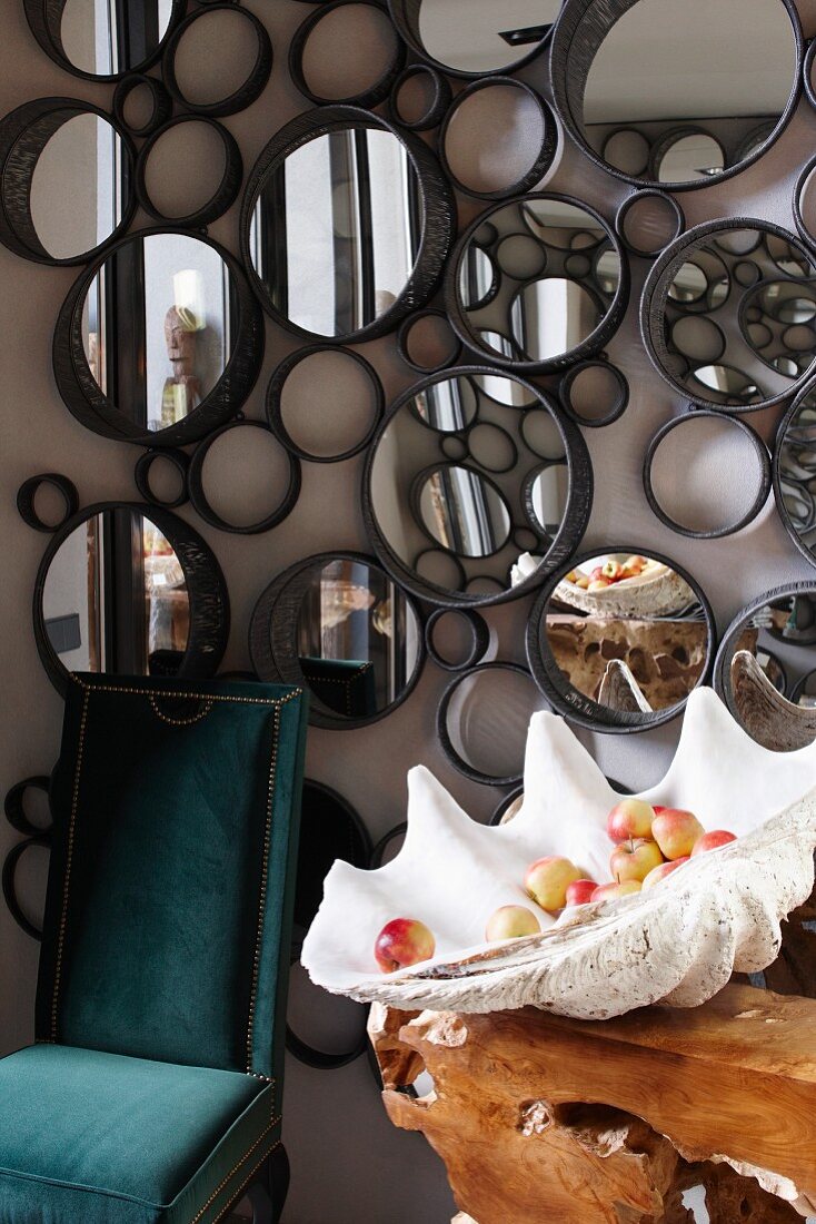 Wandobjekt aus runden Rahmen und Spiegeln, davor ein samtbezogener Antikstuhl und eine exotische Muschelschale mit Obst