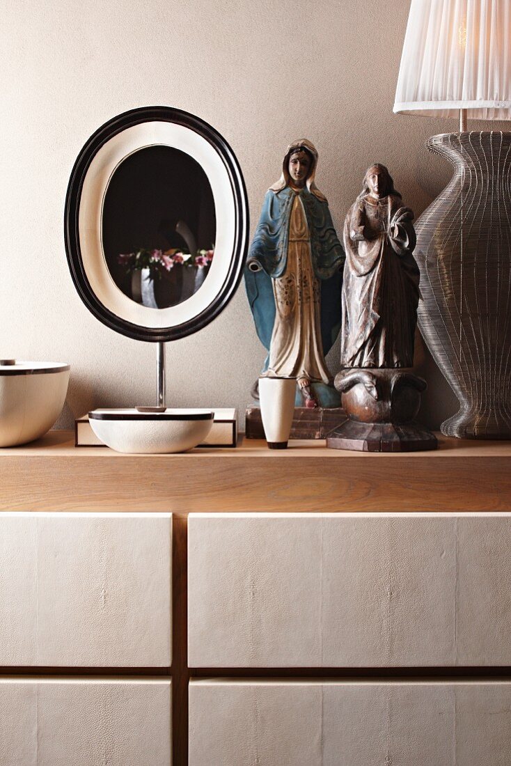 Folkloristische Frauenfigur mit Schlange und Madonnenfigur zwischen Tischleuchte und Ovalspiegel auf einer Ablage