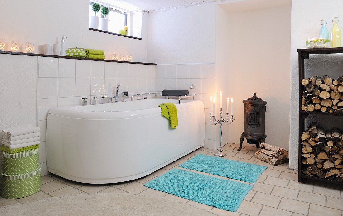 Grossräumiges Bad mit Kerzenlichtstimmung, türkise Badematten vor Badewanne, in Nische Kanonenofen und seitlich Regal mit Holzscheiten