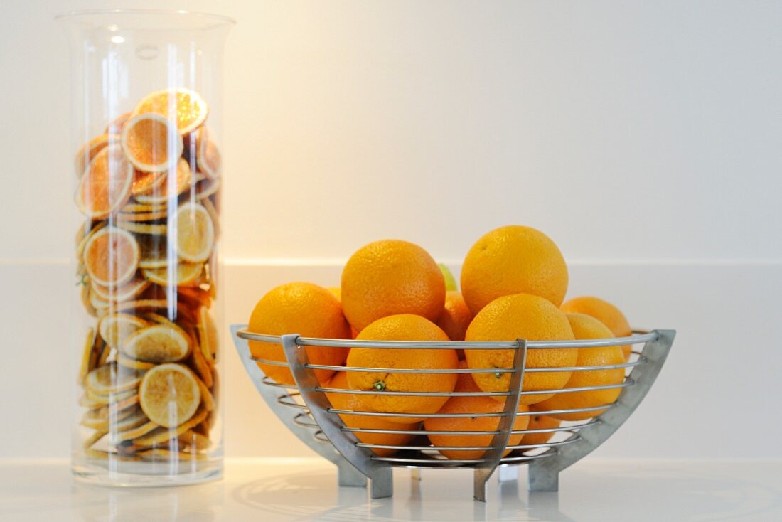 Orangen in Edelstahlschale und Orangenscheiben in zylindrischem Glasbehälter