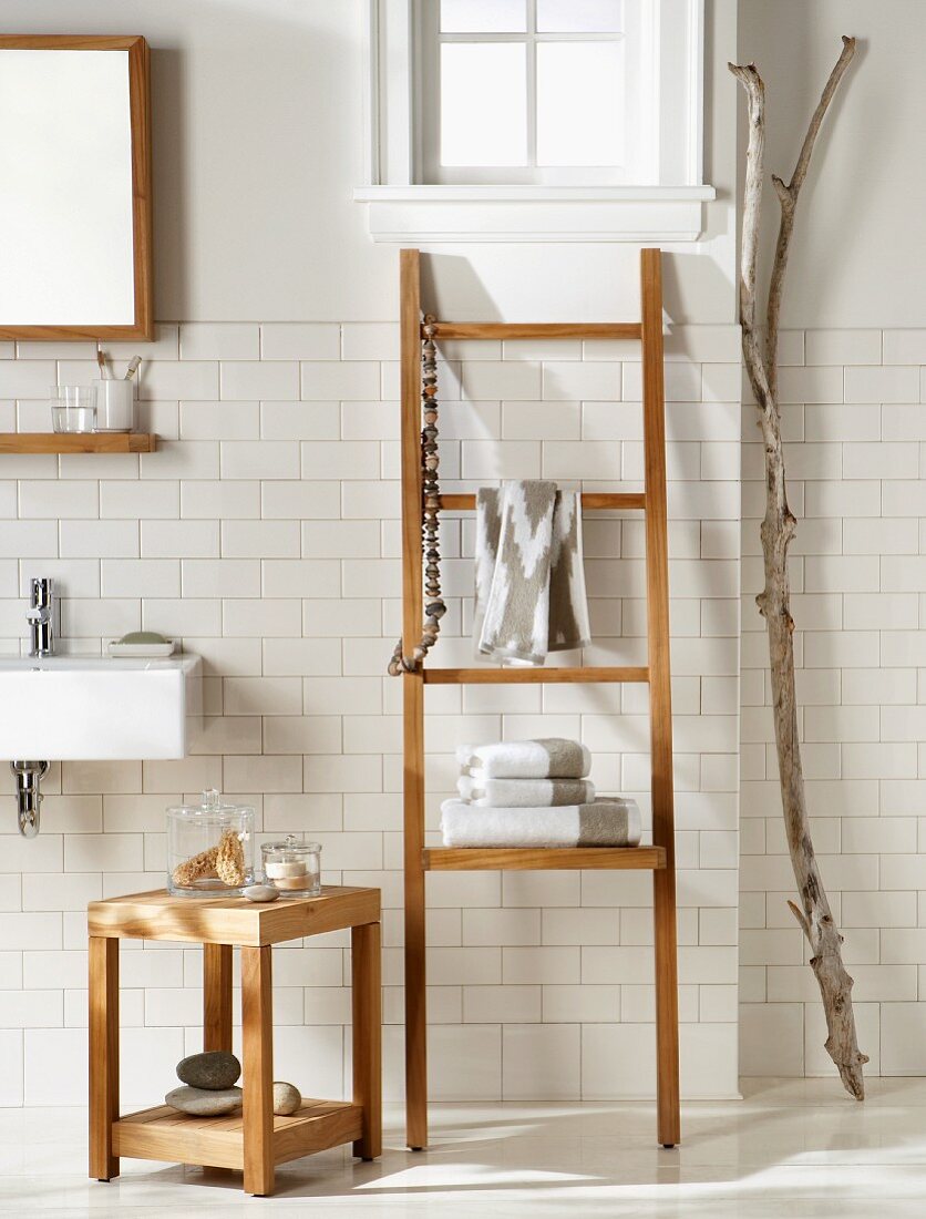 Teak stool next to ladder-style towel rack and gnarled branch against white-tiled wall in bathroom