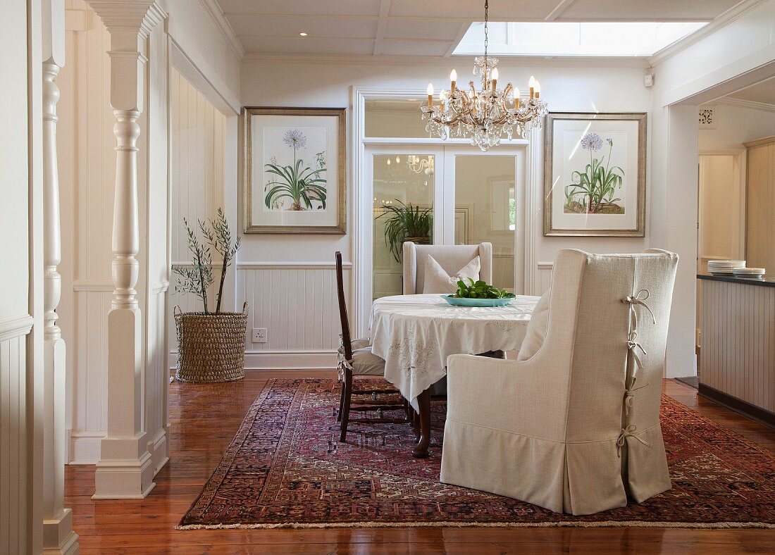 Sessel mit heller Husse und Stühle um Tisch mit Tischtuch, darüber Kronleuchter in traditionellem elegantem Esszimmer