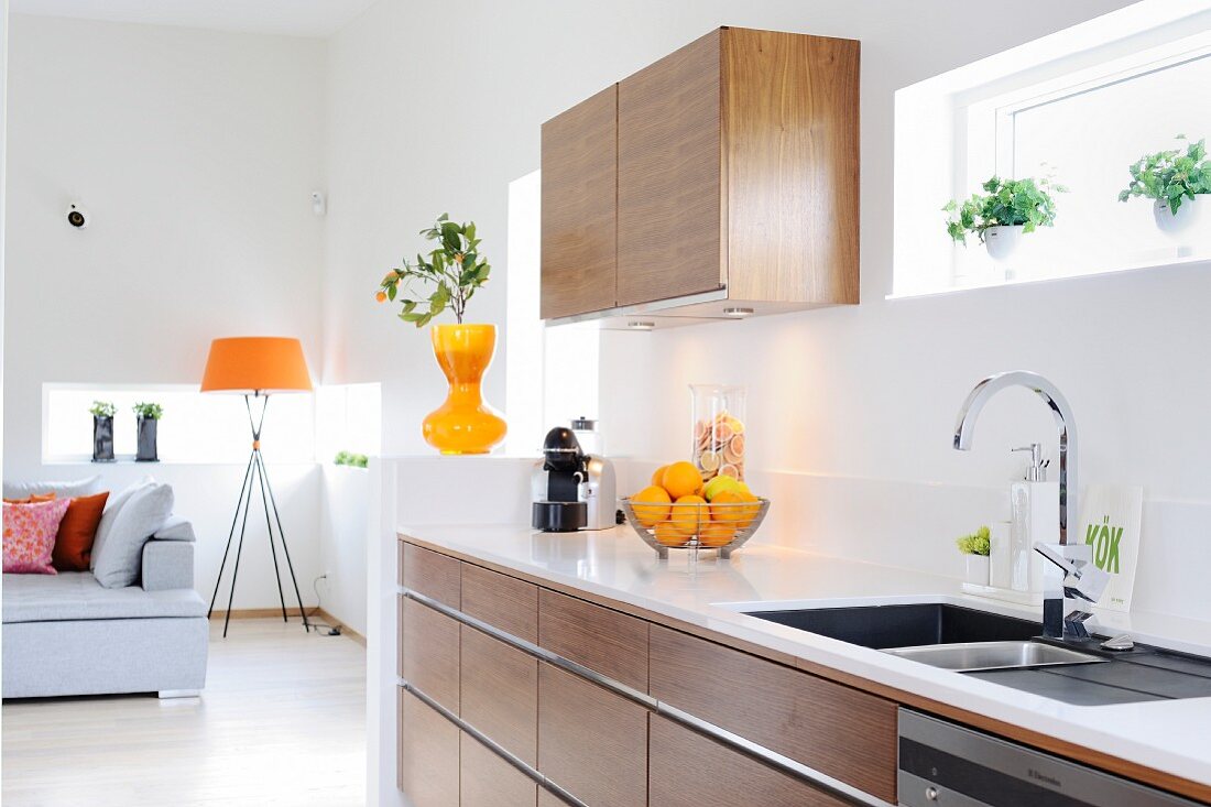 Offene Küche in Holzausführung mit weisser Arbeitsplatte, im Hintergrund Stehleuchte mit orangefarbenem Schirm in Loungebereich