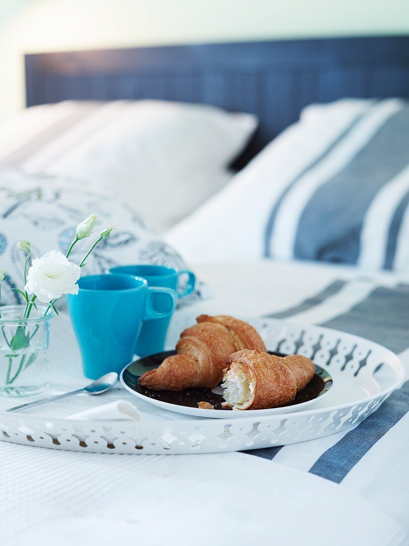 Rundes Metalltablett mit Croissant und blauen Bechern für das Frühstück im Bett
