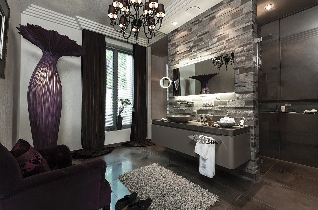 A bathroom in dark purple and green tones with a chandelier and an extravagant floor lamp, a wooden washstand and limestone against a natural stone wall