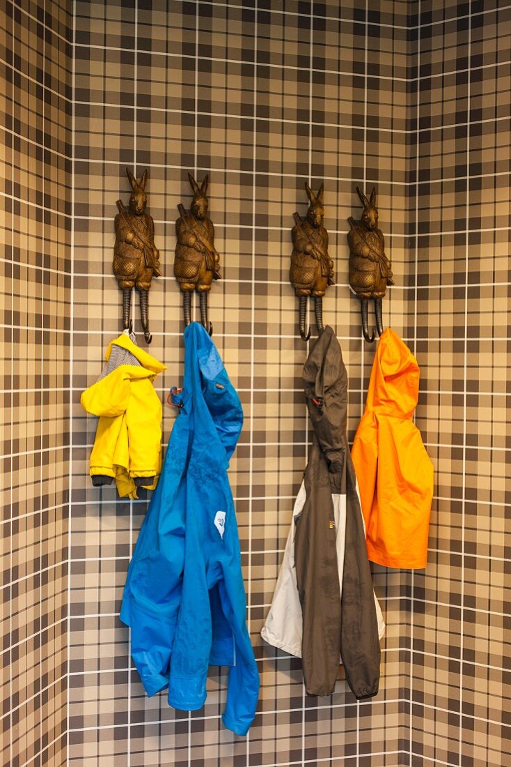 Colourful raincoats hanging on coat pegs in shape of anthropomorphic hares on brown tartan wall