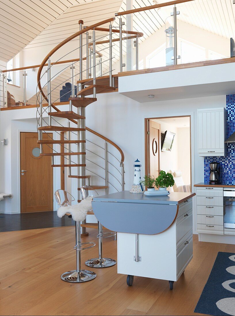 Scandinavian kitchen with island counter on castors in open-plan interior; steel and wood spiral staircase leading to upper storey