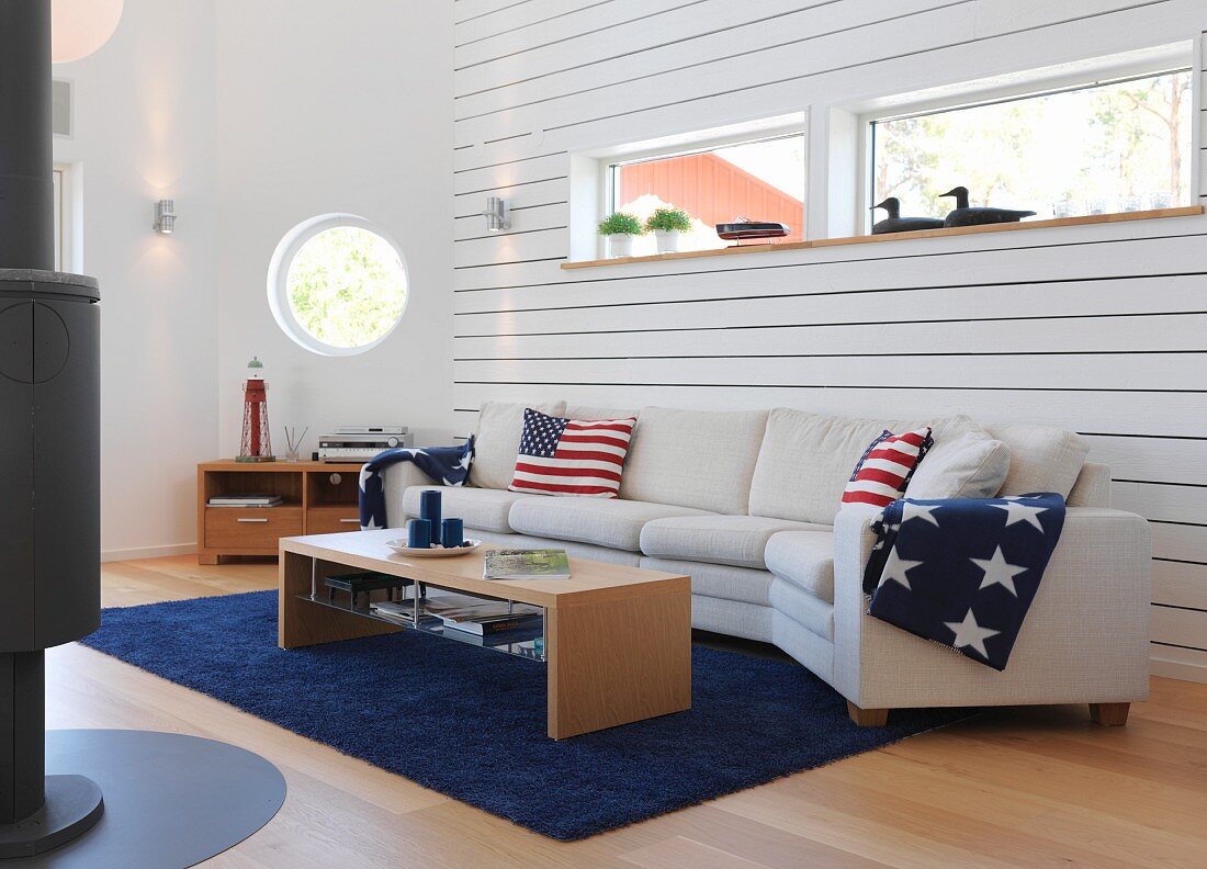 White sofa combination with stars and stripes cushions and blankets below ribbon window in wood-clad wall and porthole in background