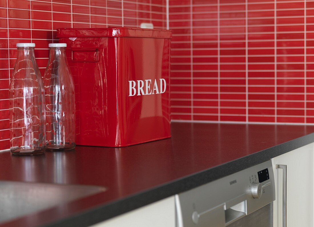 Leere Milchflaschen und Brotdose aus rotem Metall auf Küchenarbeitsfläche mit roten Mosaikfliesen als Spritzschutz