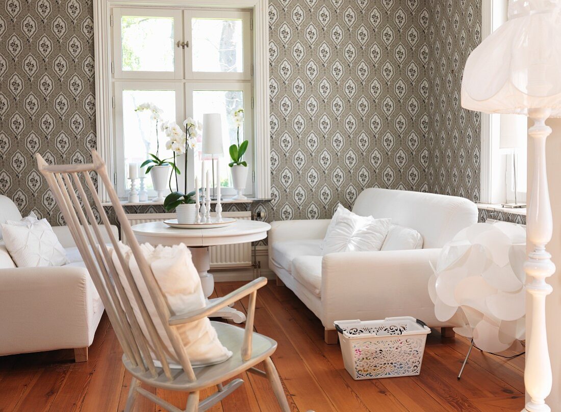 Comfortable living room with white sofa set, rocking chair, table below window and patterned wallpaper