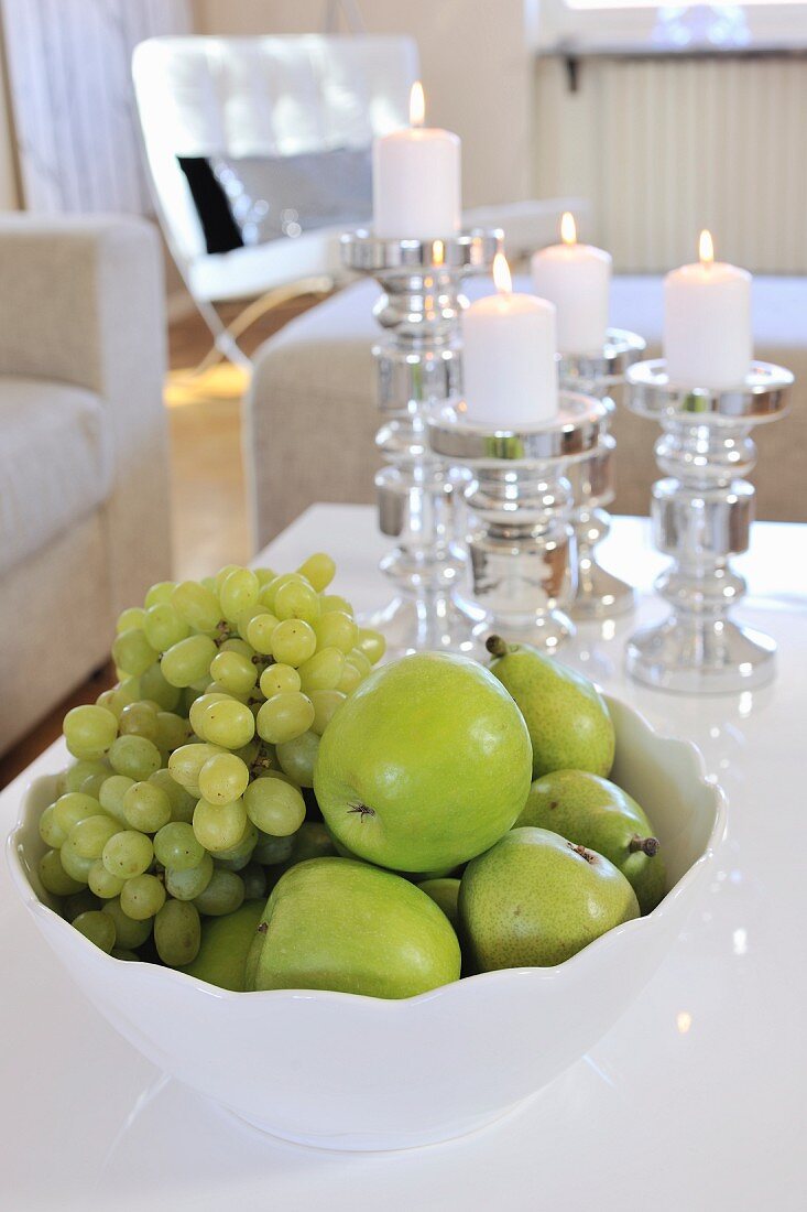Green fruit in white china bowl in front of lit candles in candlesticks