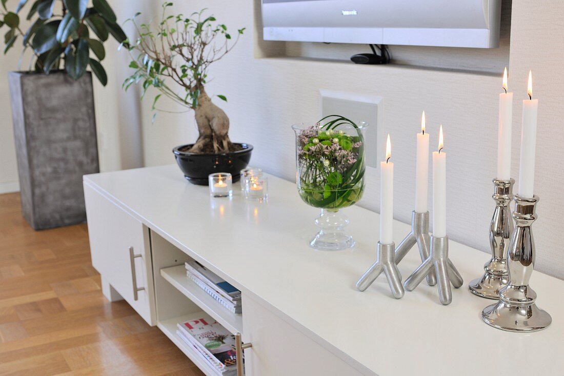 Weisses Lowboard und silberfarbene Kerzenständer mit weissen, brennenden Kerzen in modernem Wohnzimmer