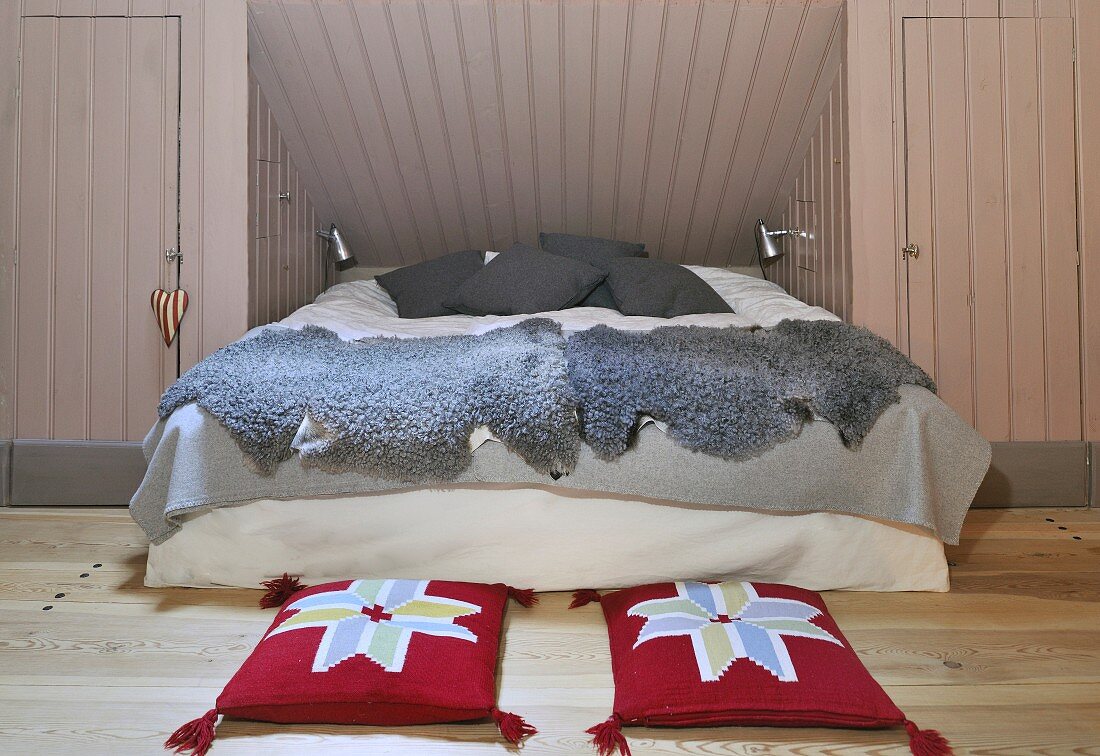 Gemütliches Bett mit grauen Tierfellen in Nische eines Dachzimmers, auf Dielenboden rote Kissen mit abstraktem Blumenmotiv