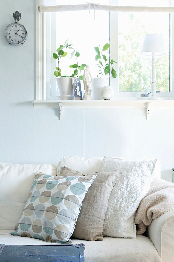 Sofa mit Kissen in verschiedenen Weissstönen vor pastellblau getönter Wand, unter Fenster