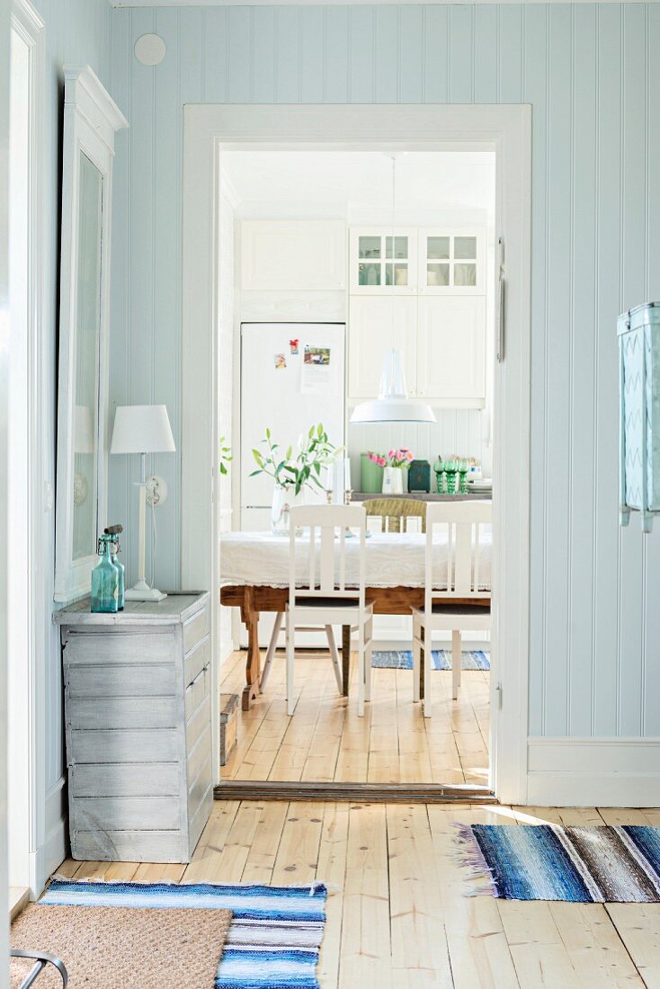 Holzverkleideter Vorraum in Pastellblau, an der Seite schlichte Kommode neben offenem Durchgang und Blick ins ländliche Esszimmer