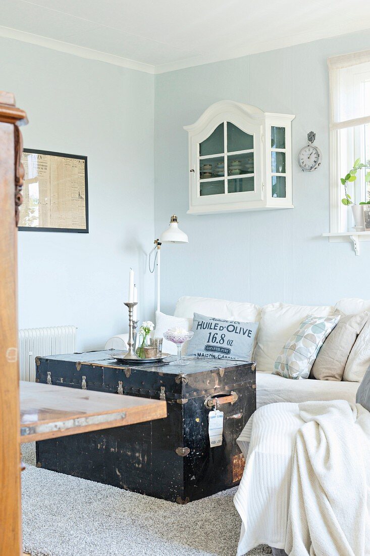 Alte Truhe vor hellem Sofa mit Kissen, darüber kleines Schränkchen in Weiß, an pastellblauer Wand aufgehängt