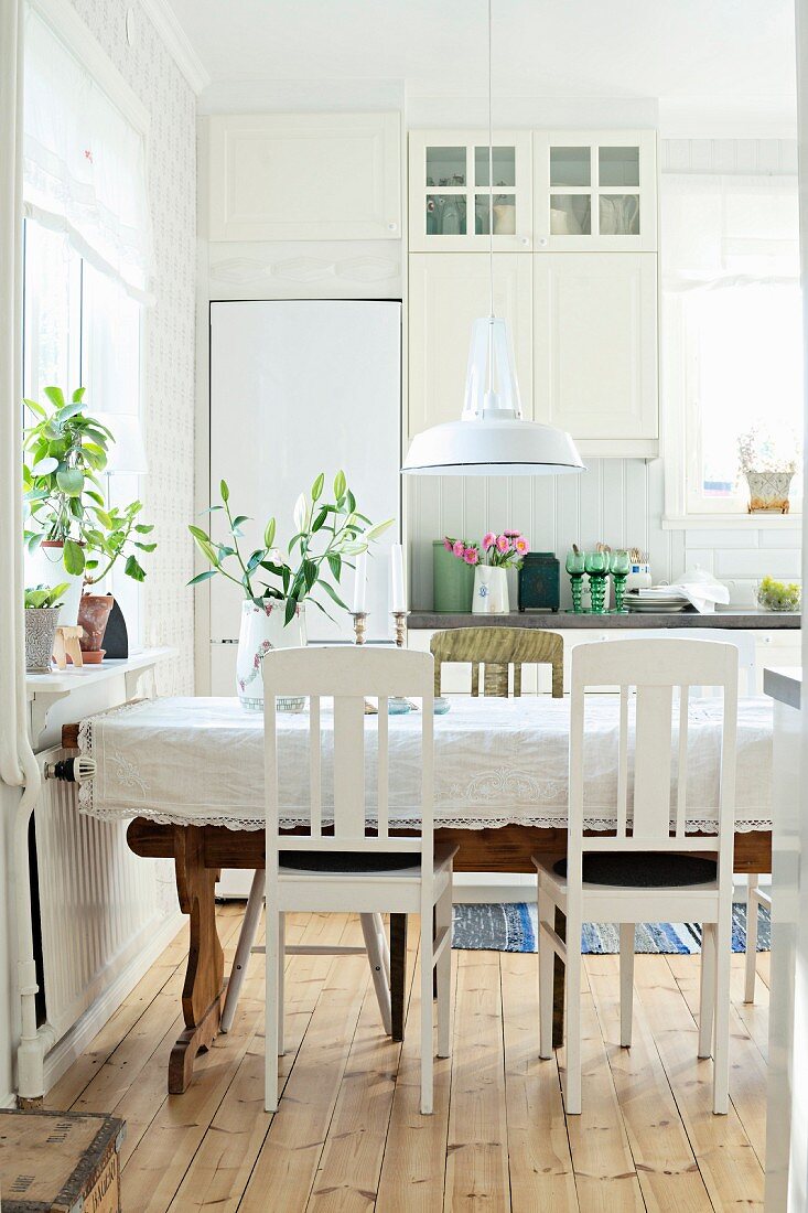 White kitchen chairs around table with tablecloth below pendant lamp with white metal lampshade in rustic dining room
