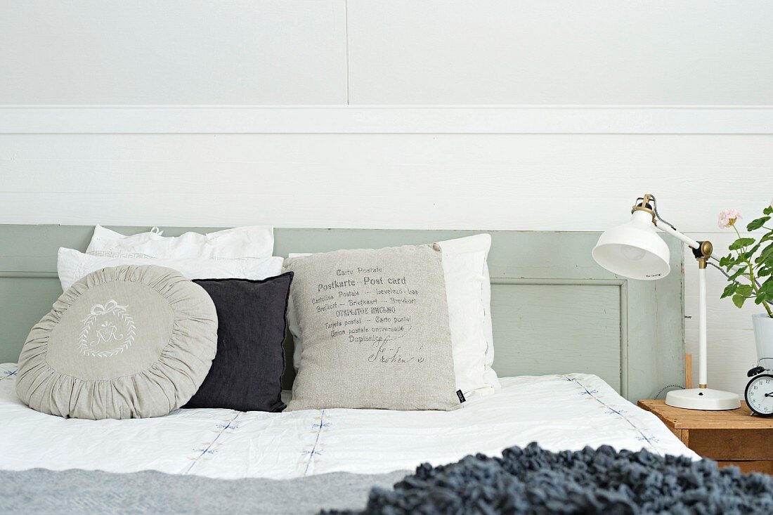 Verschiedene Kissen in Weiß- und Grautönen auf Bett, an grau lackiertem Holz Kopfteil