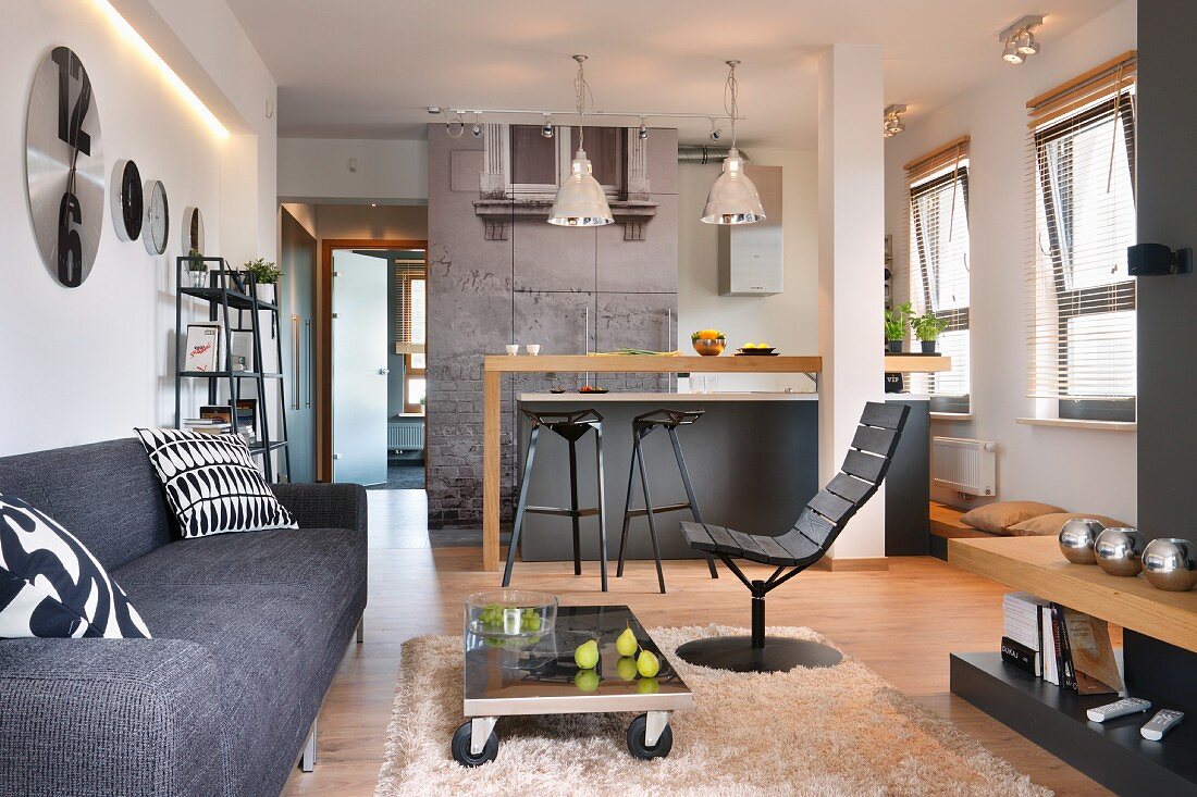 Loungebereich mit moderner, grauer Couch und rollbarem Bodentisch auf Flokatiteppich, im Hintergrund Küchentheke in offenem Wohnraum