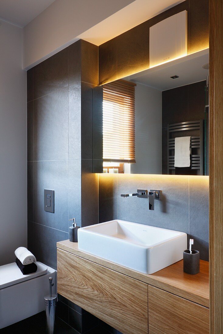 Massgefertigter Waschtisch aus Holz mit Aufbaubecken in grau gefliester Nische, oberhalb hinterleuchteter Spiegel