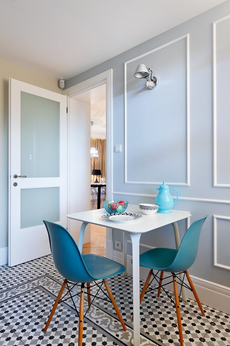 Klassikerstühle mit blau-grauer Sitzschale an weißem Frühstückstisch, vor Wand mit Paneelen aus weissen Profilleisten, geometrisches Muster auf Fliesenboden