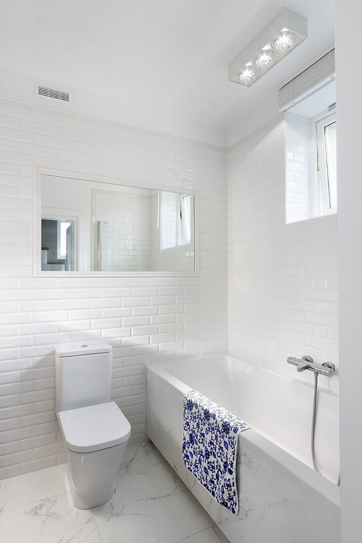 Weisses Designerbad, Toilette mit Spülkasten an gefliester Wand, neben Badewanne mit Marmorbelag an Frontseite und auf Boden