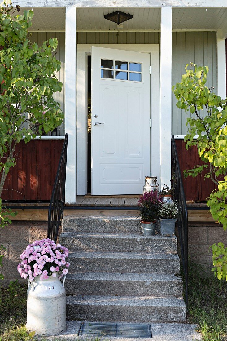 Ausschnitt eines Holzhauses mit weisser Haustür, vor Veranda Steintreppe mit Blumentöpfen, an der Seite Milchkanne mit rosa Chrysanthemen